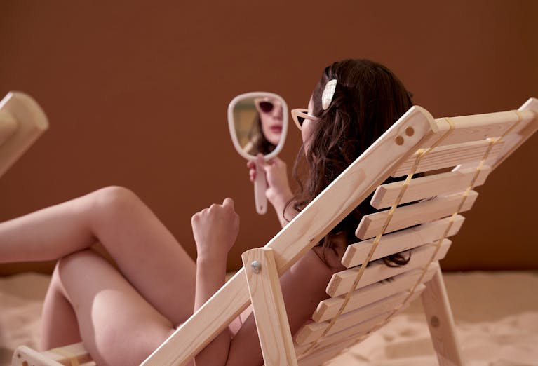 A woman in sunglasses relaxes on a deck chair holding a mirror, creating a peaceful, thoughtful scene.