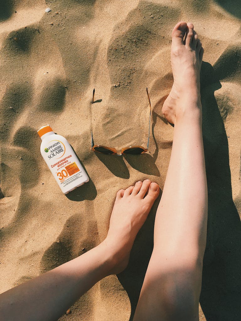 Feet on sandy beach with sunscreen bottle and sunglasses, enjoying a sunny day.