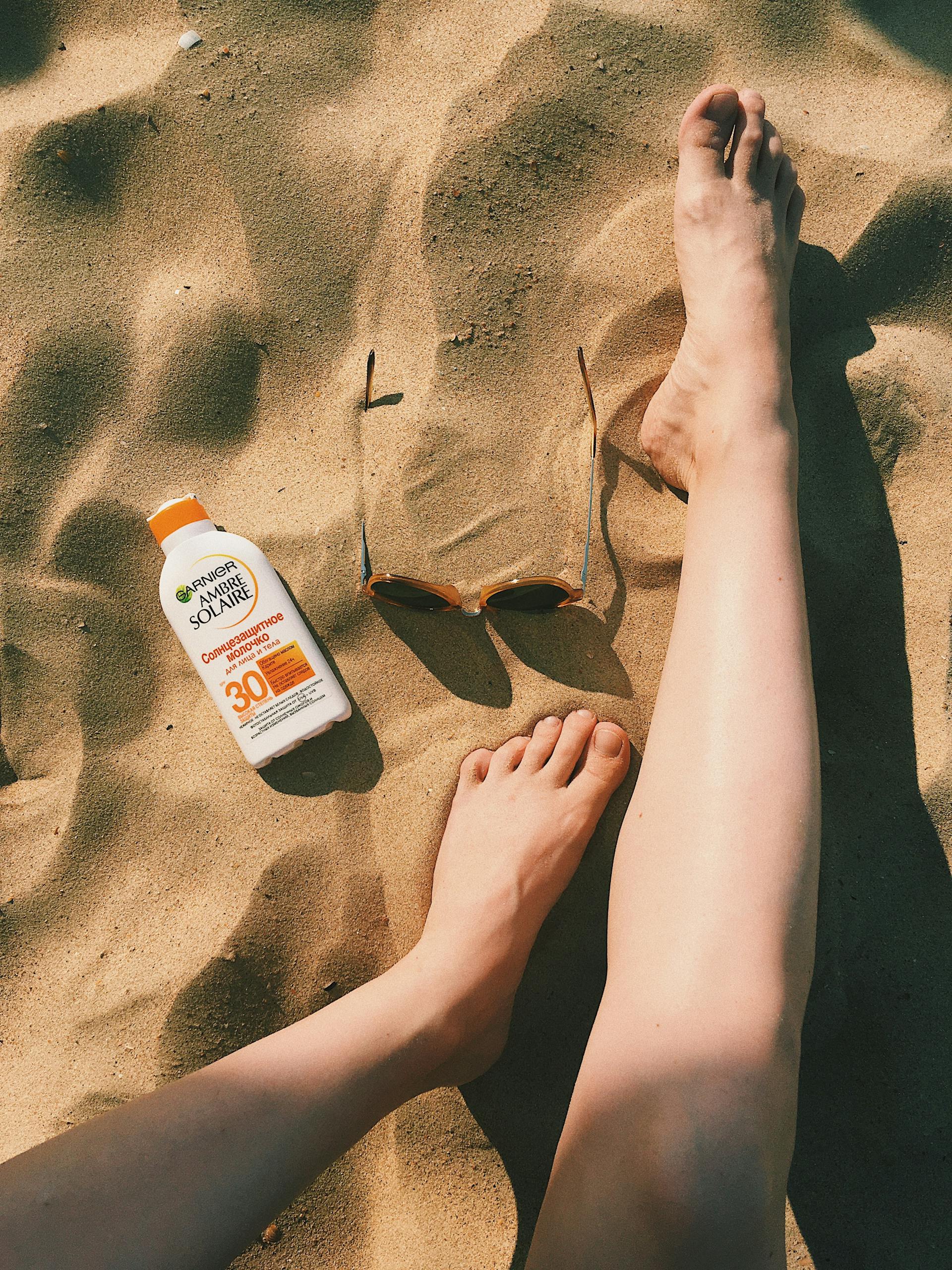 Feet on sandy beach with sunscreen bottle and sunglasses, enjoying a sunny day.