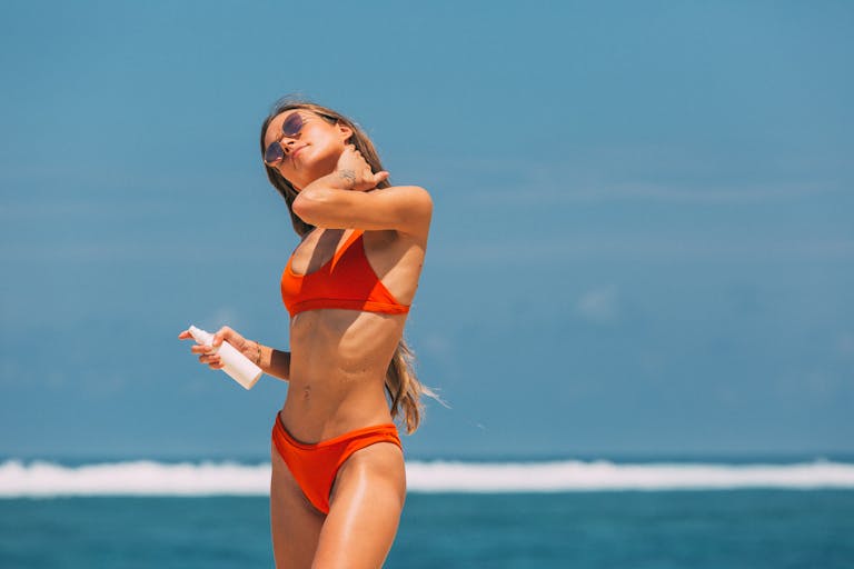 Woman in orange bikini applying sunscreen on a sunny beach day.