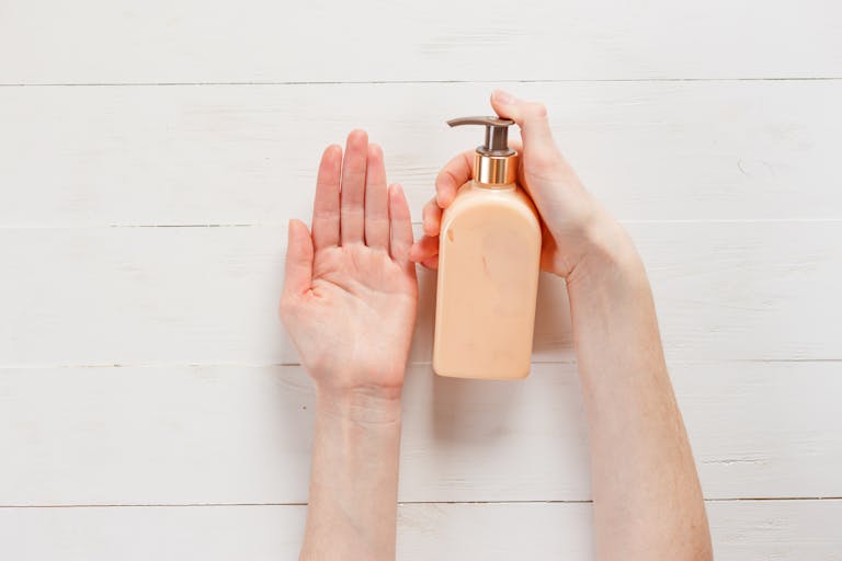 Close-up of a person's hands applying lotion using a pump bottle on a white wooden background.