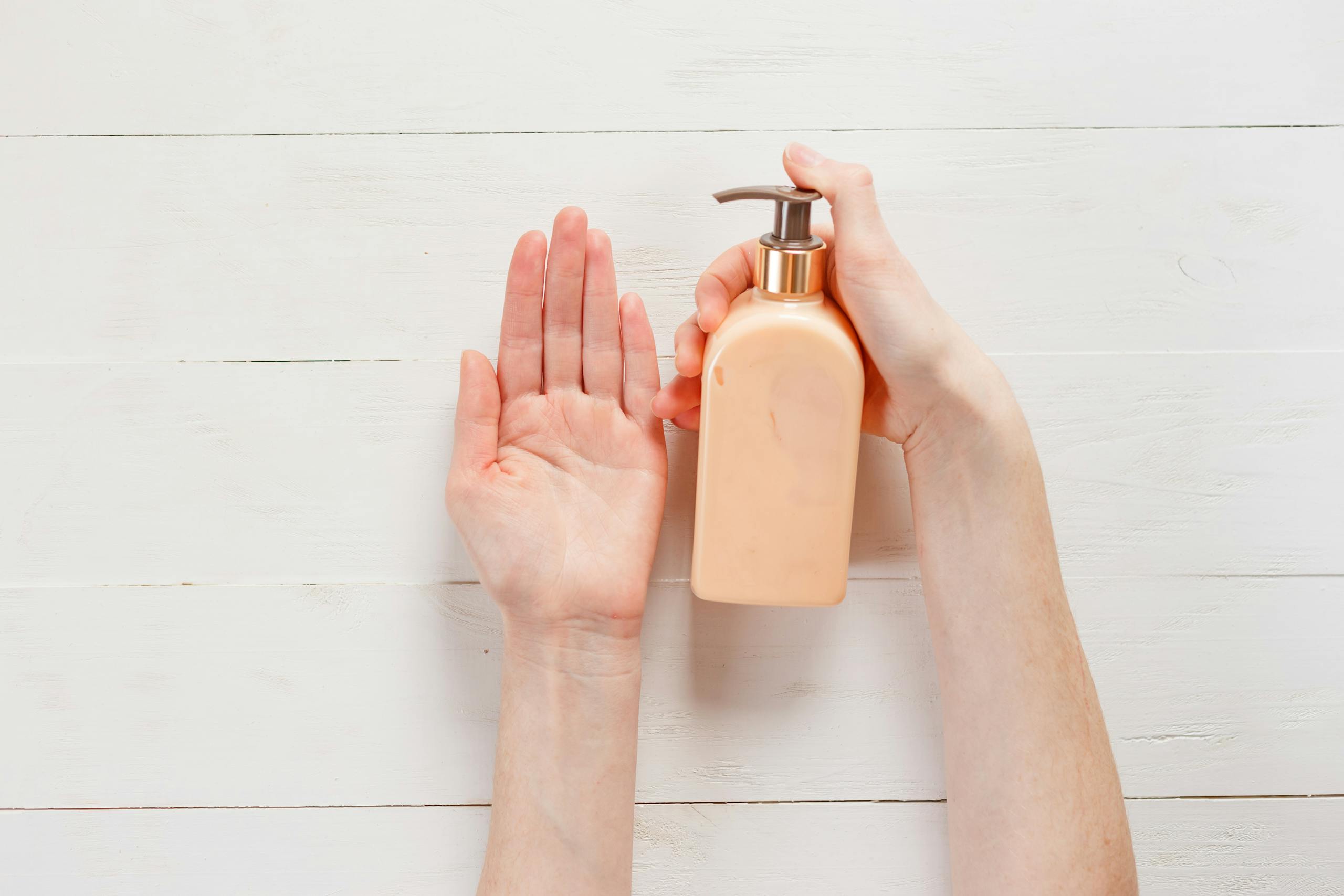 Close-up of a person's hands applying lotion using a pump bottle on a white wooden background.