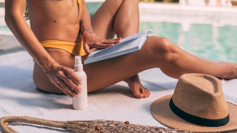 Woman in bikini reading by the pool with sunscreen and a hat, enjoying summer relaxation while tanning fast