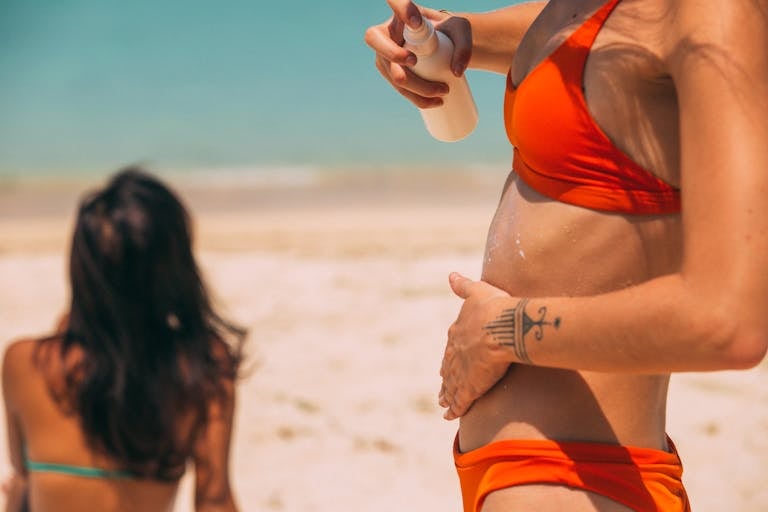 Two women on a sunny beach, one applying sunscreen and tanning lotion, both in bikinis enjoying vacation.